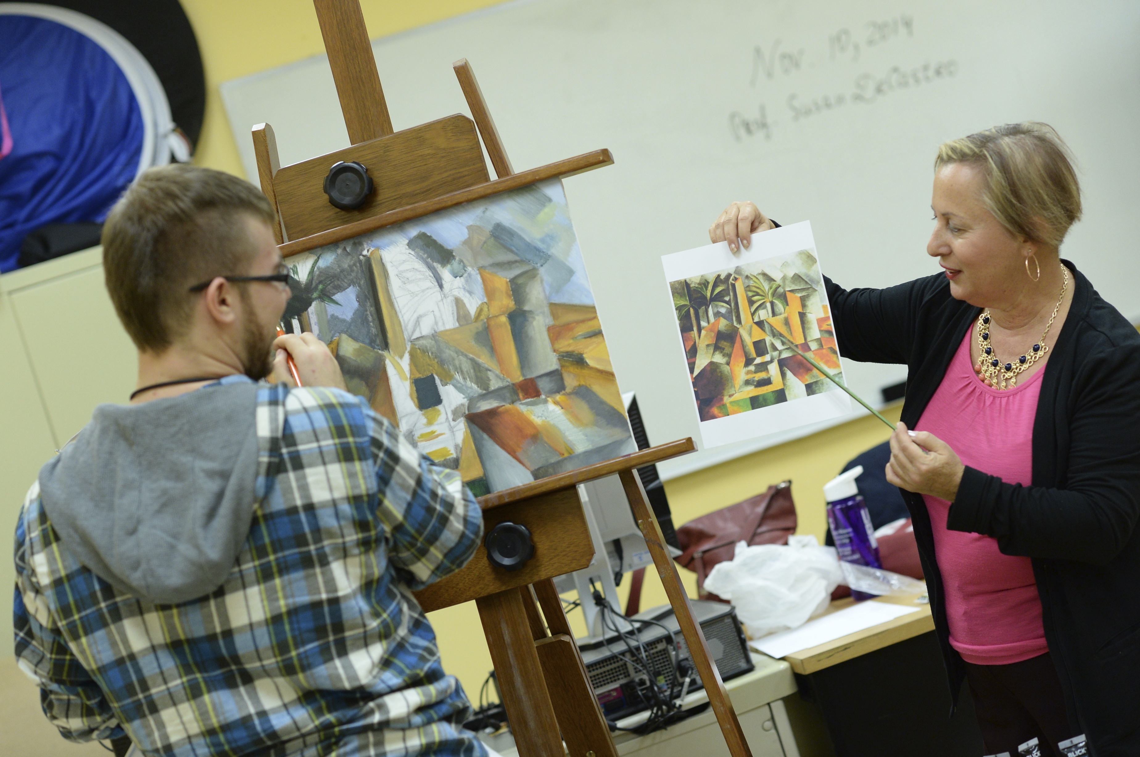 Professor Susan De Castro, holding up a print of a Picasso cubist painting, helps one of her students during class. The student is standing in front of an easel, working on his own cubist painting. 