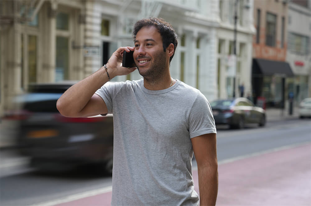 Man in grey tee shirt, on cell phone, smiling, standing in street